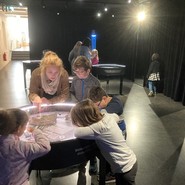 Foto von einer Lehrerin und Schüler:innen an einem Modell eines Meteoriteneinschlags (vergrößerte Bildansicht wird geöffnet)