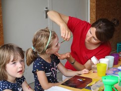 Foto einer Lehrerin, die ein Kind schminkt. Ein zweites Kind wartet. Beide Kinder sitzen auf Stühelen vor einem Tisch mit Schminkmaterialien.