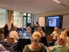Mehrere erwachsene Teilnehmende sitzen im Lehrerzimmer; zwei Referentinnen stehen vor einem Bildschirm mit einer Präsentationsfolie.