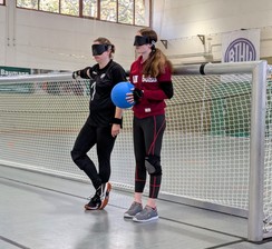 Marleen hat eine Dunkelbrille auf, hält einen Goalball in der Hand, steht mit dem Rücken am Goalballtor und neben ihr steht die Goalballnationalspielerin Annkathrin Denker.