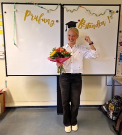 Foto von Verena Schulz mit Blumenstrauß im Lehrerzimmer vor der Aufschrift "Prüfung bestanden"
