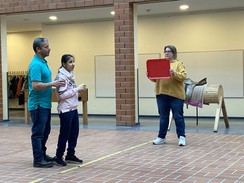 Auf dem Bild sind 3 Personen zu sehen. Ganz links steht Juan Ruiz und hält eine Schülerin an den Schultern, die vor ihm steht. Rechts steht eine Person und hält ein Tablett auf Kopfhöhe der Schülerin.