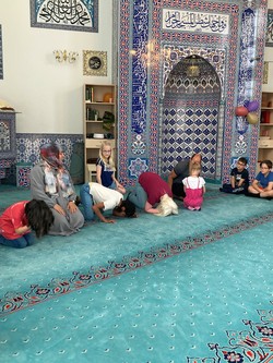 Foto von Schüler:innen und Lehrer:innen in der Gebetshaltung auf dem Teppichboden (vergrößerte Bildansicht wird geöffnet)