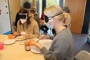 Das Bild zeigt eine Erzieherin mit Augenbinde, die ein Knäckebrot mit Marmelade bestreicht. (vergrößerte Bildansicht wird geöffnet)