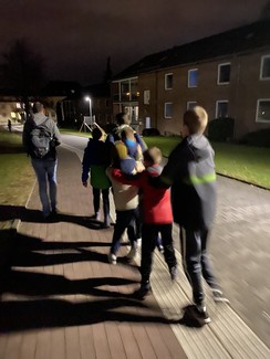 In der Abenddunkelheit laufen die 6 Spieler der Irisschulmannschaft in Alltagskleidung einen Leitstreifen eines Gehweges entlang. Sichtbar sind die Rücken der Spieler. Diese fassen mit den Händen die Schultern der voranlaufenden Spieler an. Vorweg geht eine erwachsene Person. Neben dem Weg sind zweistöckige Häuser.
