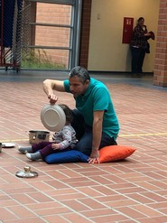 Juan Ruiz sitzt hinter einem sehr jungen Kind in der Aula der Schule auf dem Boden. Er hält ihr einen Teller vor das Gesicht. Um das Kind herum liegen Topfdeckel und Töpfe. (vergrößerte Bildansicht wird geöffnet)