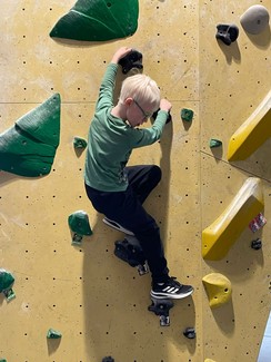 Ein Schüler klettert an einer Wand. Er steht auf zwei Griffen und hält sich ebenso mit beiden Händen fest. Der Junge schaut hinab.