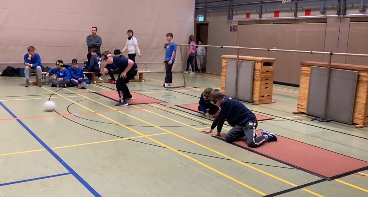 Ein Blick in eine Turnhalle mit einem Torballspielfeld. Das etwa 10 m lange Tor-Gestänge ist im Hintergrund zu sehen. Davor hocken zwei Spieler der Irisschulmannschaft in dunkelblauen T-Shirts auf Bodenmatten. Der dritte Spieler wirft den Torball stehend in einer Kegelbewegung ab. Der Ball rollt über den Boden. Am Rand des Spielfeldes sitzen 5 Spieler und steht ein Spieler einer anderen Mannschaft in hellblauen T-Shirts. 2 Kinder betreten im Hintergrund die Turnhalle. Zwei erwachsene  Personen verfolgen am Spielfeldrand das Spiel.