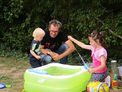 Foto von zwei Kindern mit einem Mann. Ein Kind hält eine Spielangel mit Magneten in ein Wasserbecken. Das zweite Kind schaut zu.