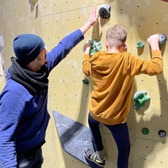 Foto: Ein Schüler klettert an einer Wand. Er steht auf zwei Griffen und hält sich ebenso mit beiden Händen fest. Ein Mann, der Trainer, steht hinter ihm und gibt ihm Tipps