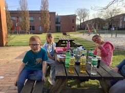 Schüler:innen sitzen an einem Picknicktisch und trinken und essen etwas. Im Hintergrund ist der Spielplatz des Schulgeländes zu sehen.