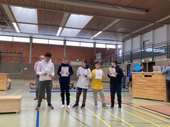 In einer Turnhalle stehen verschiedene Sportgeräte (Kästen, Matten). In der Mitte des Bildes sind 5 Spieler:innen. Sie halten Urkunden vor der Brust. Drei Spieler schauen die anderen Spieler mit Urkunde an. Zwei Spieler betrachten die Urkunde für ihre Mannschaft.