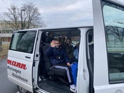 Die Fahrzeugseite eines Großraumtaxis (Bulli) füllt vollständig das Foto aus. Die Schiebetür des Fahrzeugs ist geöffnet und ein Junge schaut heraus. Er sitzt angeschnallt auf einem Sitz. Im inneren des Taxis sitzen weitere Kinder.