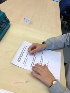 Foto von eine Teilnehmerin, die versucht tastend einen Braillebuchstaben in einer Zeile zu finden und mit einem Klebepunkt zu markieren.
