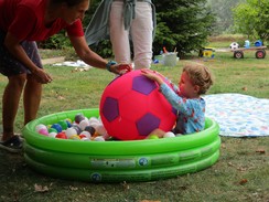 Foto eines Planschbecken mit Bällebad als "Wasser". Darin sitzt ein Kleinkind mit einem Fußball aus Kunststoff. Eine Lehrerin beugt sich zum Kind und spricht es an.