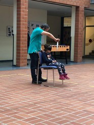 Eine Schülerin sitzt in der Aula auf einem Stuhl mit Augenbinde. Juan Ruiz hält ihr einen großen Essteller frontal vor das Gesicht. (vergrößerte Bildansicht wird geöffnet)