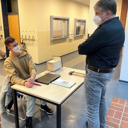 Foto von Kilian an seinem Tisch zu seinen Praktika mit Besucher (vergrößerte Bildansicht wird geöffnet)