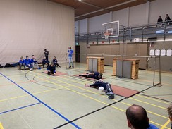 Ein Blick in eine Turnhalle mit einem Torballspielfeld. Das etwa 10 m lange Tor-Gestänge ist im Hintergrund zu sehen. Davor liegen zwei Spieler der Irisschulmannschaft in dunkelblauen T-Shirts auf Bodenmatten. Der dritte Spieler hockt auf der Matte. Der Ball rollt auf einen liegenden Spieler zu. Dieser verhindert das Weiterrollen des Balles. Am Rand des Spielfeldes sitzen 5 Spieler und steht ein Spieler einer anderen Mannschaft in hellblauen T-Shirts. Eine erwachsene  Person verfolgt am Spielfeldrand das Spiel.
